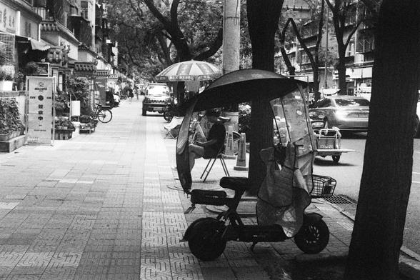 Kodak Double-X 5222 (FF)

English Alt Text:
A black-and-white image of a busy urban sidewalk. A three-wheeled electric scooter with a canopy is parked near a tree. Behind it, a person sits. Pedestrians and cyclists move along the sidewalk. Cars, including a taxi, drive on the adjacent road. Storefronts and signage line the buildings. The photo captures the rhythm of city life, blending transportation, commerce, and human activity.

中文替代文字：
一张黑白照片，描绘繁忙的城市街道。人行道旁停着一辆带遮蓬的三轮电动车。车后是一位坐着。人行道上有行人和骑车者穿…