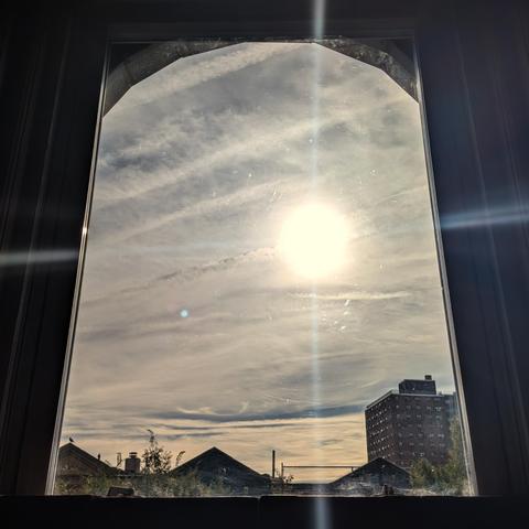 Looking through an arched window two hours and nineteen minutes after sunrise the sky is filled with complex layers of white, gray, and yellow clouds. The sun is above the houses, to the right of center, creating a lens flare which looks like cross hairs across the frame. Pointed roofs of Harlem brownstones are silhouetted across the street, and a taller apartment building can be seen in the distance. The green tops of two trees are on the bottom and right. The window is grimy on the bottom.