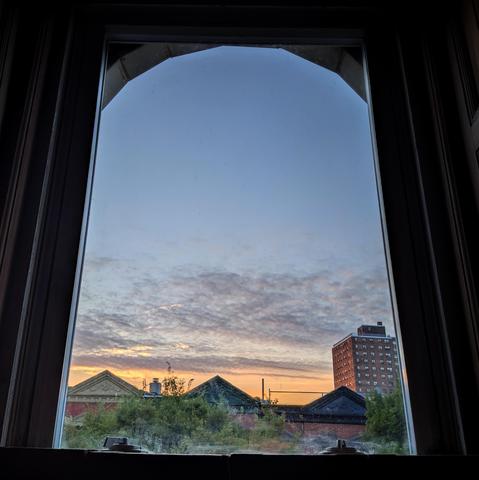 Looking through an arched window thirteen minutes after sunrise the sky fades from pale orange at the horizon to dusty blue up above, with a band of gray clouds in the lower and middle sky. Pointed roofs of Harlem brownstones are silhouetted across the street, and a taller apartment building can be seen in the distance. The green tops of two trees are on the bottom and right. 