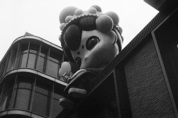 Kodak Double-X 5222 (FF)

English Alt Text:
A whimsical sculpture of a cartoon-like figure is mounted above the entrance of a modern building. The figure has a round head, oversized eyes, and spherical decorations resembling hair or ornaments. The building features curved glass windows and brick walls, blending playful art with contemporary architecture. The sculpture’s exaggerated proportions and joyful expression make it a focal point, inviting curiosity and amusement.

中文替代文本：
一座卡通风格的雕塑安装在现代…