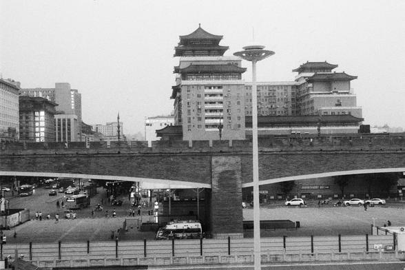 Kodak Double-X 5222 (FF)

English Alt Text:
A black-and-white cityscape showing traditional Chinese buildings with ornate, curved roofs in the background. These structures resemble temples or historic halls. In the foreground, a large stone or concrete bridge with arches spans the scene. Beneath the bridge, cars are parked and pedestrians walk, suggesting a busy urban setting. Streetlights and modern buildings add contrast. The image captures the coexistence of ancient architecture and modern l…