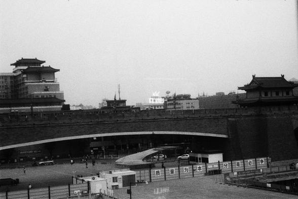 Kodak Double-X 5222 (FF)

English Alt Text:
A black-and-white urban scene featuring a large stone bridge or elevated roadway spanning the center. Below, a plaza with people walking and vehicles parked. Traditional East Asian buildings with tiered roofs line the area, contrasting with modern buildings and a communications tower in the background. A fence in the foreground displays Chinese characters. The image captures cultural contrast and active public space.

中文替代文字：
这是一张黑白城市照片，画面中央是一座大型石桥或高架…