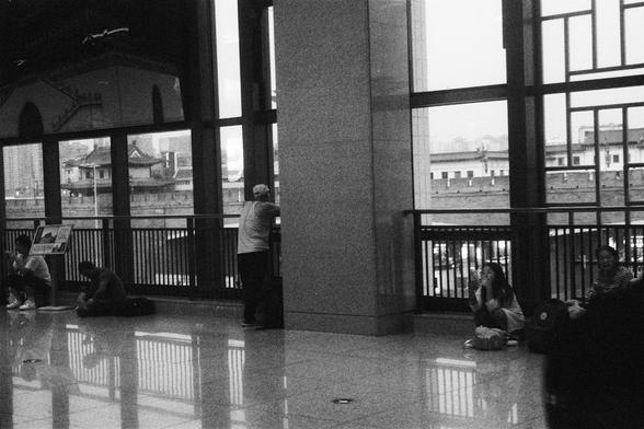 Kodak Double-X 5222 (FF)

English Alt Text:
A black-and-white photo taken inside a large building with tall glass windows. Outside, traditional-style architecture is visible. Inside, people sit or stand near the windows. One person leans against a column, gazing out. Others sit on the floor, some chatting or using phones. The reflective floor adds depth. The scene contrasts modern interior design with historical exterior elements, evoking quiet observation and cultural reflection.

中文替代文字：
这是一张…