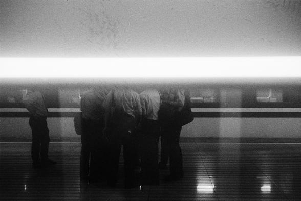 Kodak Double-X 5222 (FF)

English Alt Text:
A black-and-white photo of a platform. A group of people stands near the edge, partially obscured by a bright horizontal light streak across the middle of the image. The light creates a dramatic effect, hiding the upper bodies and faces of the individuals. Some appear to be talking, others waiting. A train is visible in the background with horizontal stripes and windows. The platform floor has a striped pattern, adding geometric contrast. The overall …