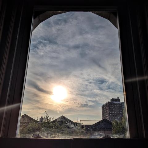 Looking through an arched window eighty minutes after sunrise the sky is filled with complex layers of white, gray, and yellow clouds. The sun is above the houses, slightly to the left of center, and creating a lens flare as a curved horizontal white line across the frame. Pointed roofs of Harlem brownstones are silhouetted across the street, and a taller apartment building can be seen in the distance. The tops of two trees are on the bottom and right. The window is grimy on the bottom.