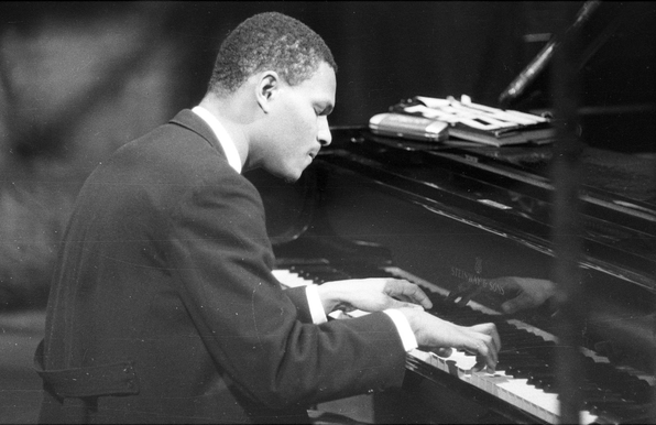 A youthful McCoy Tyner is heavily concentrating as he's playing a grand piano. The picture is black and white. Tyner is wearing a black coat over a dress shirt. 