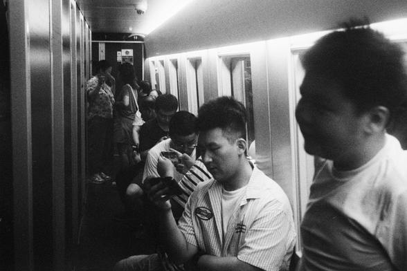 Kodak Double-X 5222 (FF)

English Alt Text:
A black-and-white photo captures a narrow corridor inside a train. Several people are seated along the wall, each absorbed in their smartphones. Others stand or walk through the passage, creating a sense of motion. The lighting is dim, with a single strip of light running overhead, casting soft shadows and reflections on the metallic surfaces. The atmosphere feels quiet and introspective, highlighting the solitude often found in public spaces. The ima…
