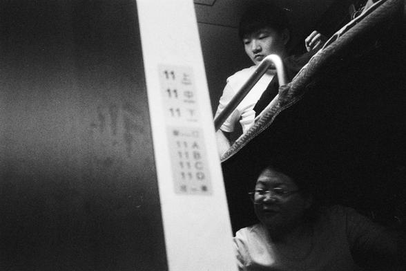 Kodak Double-X 5222 (FF)

English Alt Text:
Inside a train compartment with bunk beds, a black-and-white photo shows two passengers. One sits on the lower bunk, gazing sideways with a calm expression. The other is partially visible on the upper bunk, looking downward through a metal railing. A vertical panel nearby displays berth numbers in Chinese characters and digits. The lighting is dim and grainy, giving the image a candid, documentary feel. The scene suggests quiet moments during overnigh…