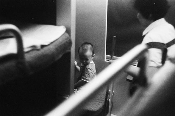 Kodak Double-X 5222 (FF)

English Alt Text:
A black-and-white photo captures a young child sitting quietly near a wall, possibly inside a train or bus. The child looks downward, with a thoughtful or tired expression. An adult stands nearby, partially visible on the right side. The foreground includes railings and seat edges, framing the child and adding depth. The soft lighting and candid composition evoke a sense of intimacy and quiet observation, suggesting a moment of rest or contemplation d…