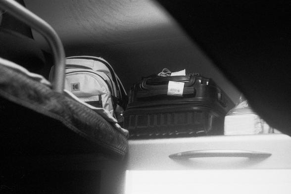 Kodak Double-X 5222 (FF)

English Alt Text:
A black-and-white photo shows an overhead luggage shelf inside a train or bus. On the shelf are a light-colored backpack with a logo, a black hard-shell suitcase with a luggage tag, and a white box or package. Below the shelf, part of a seat or sleeping berth is visible. The image is taken from a low angle, emphasizing the compact storage space. The scene reflects the practical side of travel, focusing on personal belongings and the confined nature of…