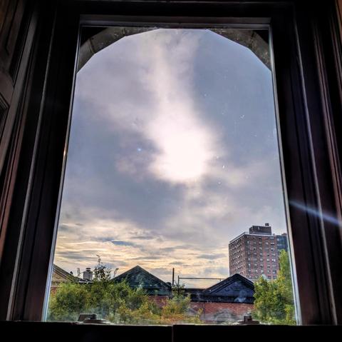 Looking through an arched window two hours  after sunrise the sky is filled with complex layers of white, gray, and yellow clouds. The sun is a blur in the middle of the sky, creating a lens flare as a diagonal white line on the right. Pointed roofs of Harlem brownstones with red brickwork are across the street, and a taller apartment building can be seen in the distance. The green tops of two trees are on the bottom and right. The window is grimy on the bottom.