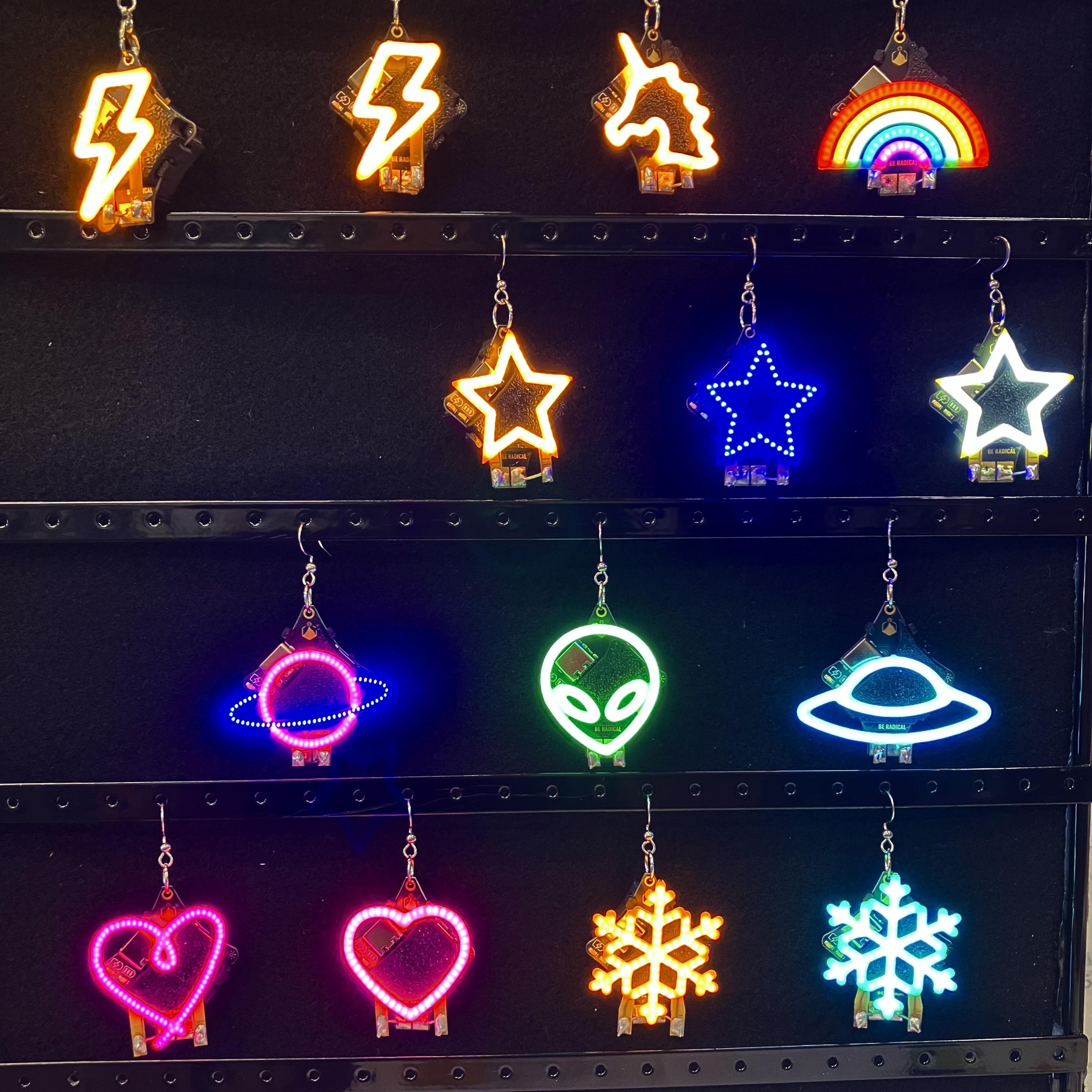 Glowies are displayed hanging from push earrings against a dark background. Featured our two lightning bolts, a unicorn, a rainbow three stars, a planet and alien a UFO two hearts and two snowflakes.