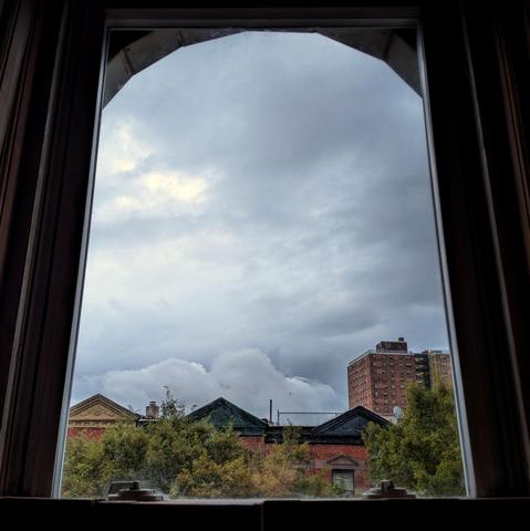 Looking through an arched window half an hour after sunrise the sky is full of blue-grey rain clouds. Pointed roofs of Harlem brownstones with red brickwork are across the street, and a taller apartment building can be seen in the distance. The green tops of two trees are on the bottom and right.