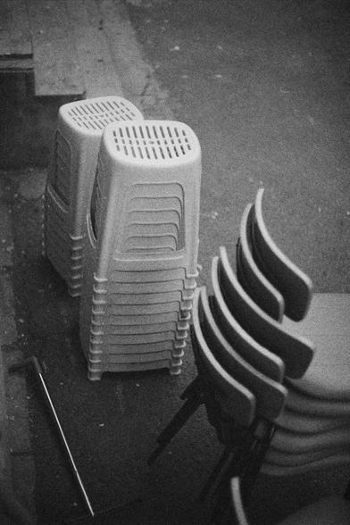 Ilford HP5 Plus 400 (FF)

English Alt Text:
A black-and-white photo of outdoor furniture arranged in neat stacks. On the left, two tall columns of plastic stools are nested together, each stool featuring small ventilation holes on top. On the right, a stack of plastic chairs with curved backrests and metal legs leans slightly. The chairs are also nested, forming a compact tower. The ground is paved, and a metal rod lies nearby. In the upper left corner, wooden steps suggest proximity to a build…