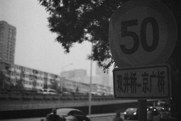 Ilford HP5 Plus 400 (FF)

English Alt Text:
A black-and-white photo of a city street scene. In the foreground, a circular traffic sign displays the number “50,” indicating a speed limit of 50 km/h. Below it, a rectangular sign with Chinese characters reads “Shuangjing Bridge – Jingguang Bridge.” The signs are mounted on a pole near a leafy tree. In the background, cars and a motorcycle move along a wide road flanked by buildings, including a high-rise and a long residential block. The grainy te…