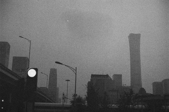 Ilford HP5 Plus 400 (FF)

English Alt Text:
A monochrome photo of a city skyline shrouded in smog or fog. Several tall buildings are visible, including a striking skyscraper shaped like a twisted column. A green traffic light glows in the foreground, mounted on a pole near streetlights and an overpass. The air appears thick, reducing visibility and softening the outlines of buildings. The image evokes urban density and environmental tension, with a quiet, surreal mood.

中文替代文本：
这是一张黑白城市天际线照片，画面…