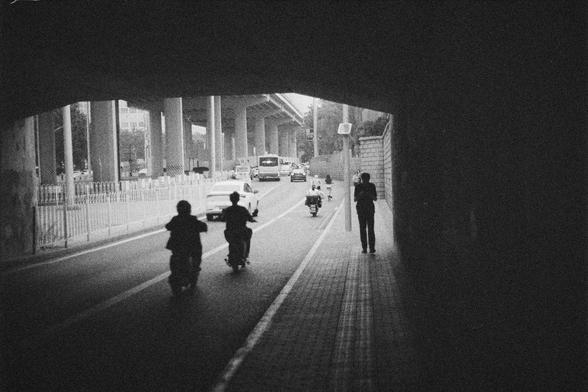 Ilford HP5 Plus 400 (FF)

English Alt Text:
A black-and-white photo taken from inside a tunnel or underpass, looking out onto a busy urban road. Several people ride scooters or motorcycles, while one person walks along the sidewalk. The road continues under a series of elevated structures supported by thick columns. In the distance, a bus and a car are visible. The tunnel’s shadow frames the scene, creating strong contrasts between light and dark. The photo captures a moment of city life in mot…