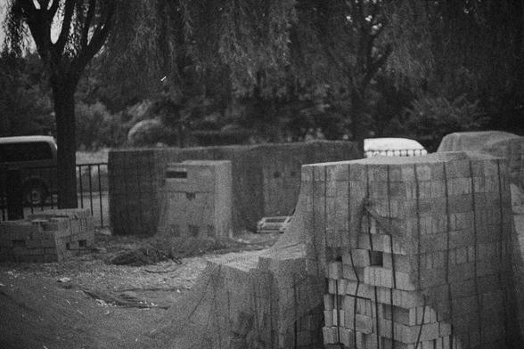 Ilford HP5 Plus 400 (FF)

English Alt Text:
A black-and-white image of an outdoor construction or storage site. Stacks of bricks and concrete blocks dominate the foreground, some covered with tarps. Behind them, a fence encloses the area, with trees and a partially visible vehicle beyond. The grainy texture and muted tones give the scene a raw, industrial, and slightly nostalgic atmosphere.

中文替代文字：
一张黑白照片，展示一个户外施工或储存场地。前景是堆叠的砖块和混凝土块，有些覆盖着防水布。背景中有围栏，围栏外是树木和一辆部分可见的车辆。颗粒感和灰暗色调营造出粗犷的工业氛围，略带怀旧气息。