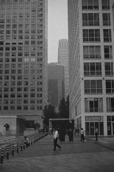 Ilford HP5 Plus 400 (FF)

English Alt Text:
A bustling urban walkway flanked by tall office buildings with grid-like glass facades. People walk or sit along the path, some near bollards that separate the walkway from the road. In the distance, a skyscraper with a crown-like top rises prominently. Signs in English and Asian characters are visible. The black-and-white palette gives the scene a timeless, documentary feel.

中文替代文字：
一条繁忙的城市步道，两侧是高大的办公楼，玻璃幕墙呈网格状。行人沿路行走或坐在路边，有些靠近分隔道路的柱墩。远处一座顶部如皇冠般的摩天大…