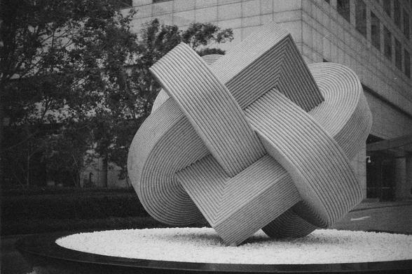 Ilford HP5 Plus 400 (FF)

English Alt Text:
A striking abstract sculpture made of stone stands outdoors in a modern urban setting. The sculpture features interlocking curved and rectangular shapes with ribbed textures, forming a complex geometric design. It rests on a circular base covered in white pebbles. Surrounding the sculpture are trees and sleek buildings, suggesting a public art installation. The interplay of shapes and textures invites interpretation, evoking thoughts of movement, bala…