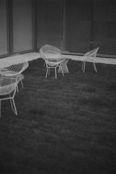 Ilford HP5 Plus 400 (FF)

English Alt Text:
A black-and-white photo of a quiet outdoor patio. Several wireframe chairs and a matching table sit on a grassy surface. The furniture is made of thin metal rods forming a mesh pattern, giving it a light, airy appearance. Behind the seating area is a modern building with large glass panels and vertical blinds, reflecting soft light. The composition is minimalistic, with clean lines and a mid-century modern aesthetic. Shadows from the furniture and bui…