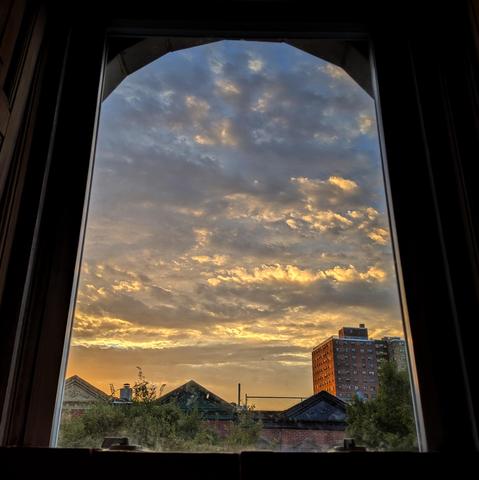 Looking through an arched window eleven minutes after sunrise the sky fades from orange at the horizon to dusty blue up above. Complex layers of clouds are underlit by the rising sun, turning yellow. Pointed roofs of Harlem brownstones are silhouetted across the street, and a taller apartment building can be seen in the distance. The green tops of two trees are on the bottom and right.