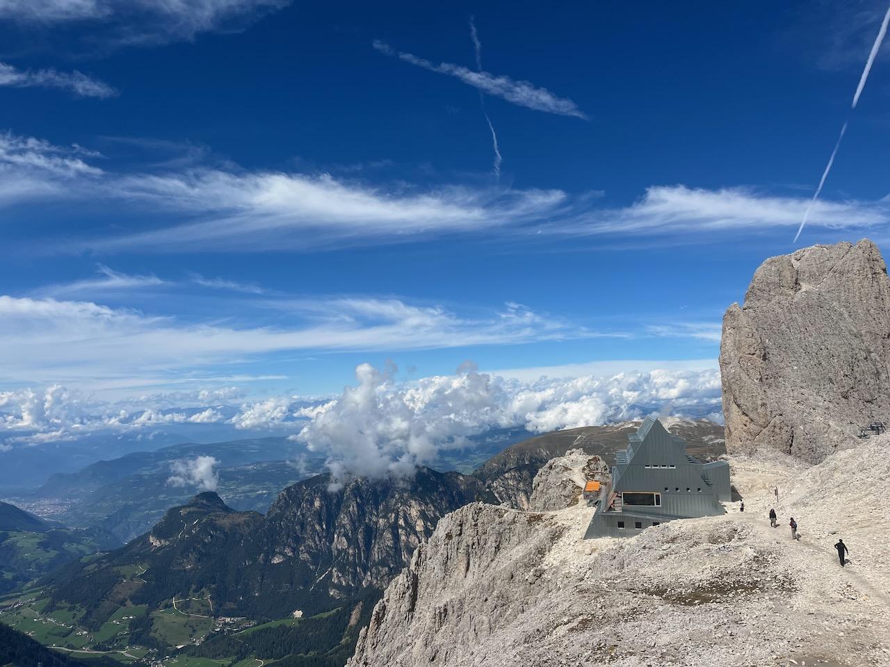 A side view on Rifugio Santner, a triangular prism lying on its side clad in aluminium. Far below, a green valley can be seen. The sky above is intensely blue with some high white clouds.