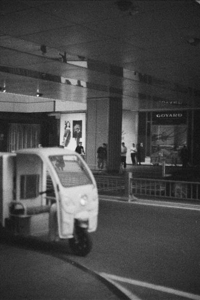 Ilford HP5 Plus 400 (FF)

English Alt Text:
A small three-wheeled electric vehicle is parked in the foreground, slightly out of focus. In the background, people walk past luxury storefronts including one labeled “GOYARD.” The black-and-white photo emphasizes the contrast between mobility and commerce.

中文替代文字：
前景是一辆小型三轮电动车，略微模糊。背景中人们在奢侈品店前走动，其中一家店标为“GOYARD”。黑白照片突出了交通工具与商业空间的对比。