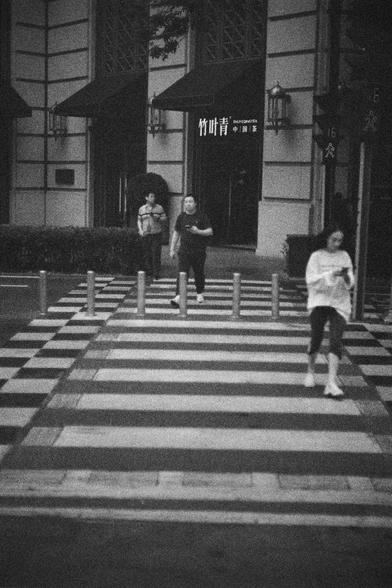 Ilford HP5 Plus 400 (FF)

English Alt Text:
Three people walk across a zebra-striped crosswalk bordered by short metal posts. Behind them is a building with large windows and awnings. A sign in Chinese reads “Bamboo Leaf Green” and “Chinese Tea.” The photo has a grainy, film-like texture.

中文替代文字：
三人正走在斑马线人行道上，周围有矮金属柱围栏。背景是一栋带有大窗户和遮阳篷的建筑，招牌上写着“竹叶青”和“中国茶”。照片呈颗粒状，仿佛胶片拍摄。