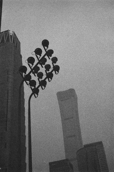Ilford HP5 Plus 400 (FF)

English Alt Text:
A black-and-white cityscape featuring a tall streetlight in the foreground with multiple lamps arranged in a grid. Behind it, modern skyscrapers rise, including one with a curved, futuristic design and another with a traditional rectangular shape. The grainy texture adds a vintage or artistic feel.

中文替代文字：
一幅黑白城市风景照，前景是一盏高大的路灯，灯头呈网格状排列。背景中有几座高楼，其中一座是曲线造型的现代摩天大楼，另一座则是传统的矩形建筑。画面有颗粒感，呈现出复古或艺术风格。