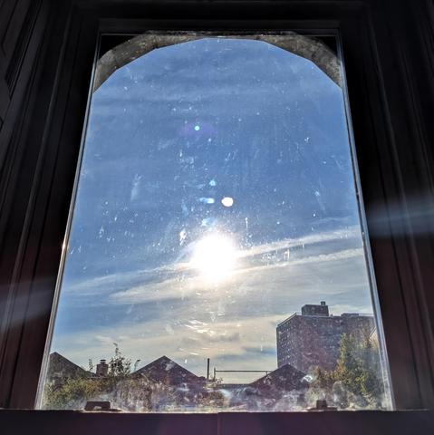 Looking through an arched window 104 minutes after sunrise the sun is in the middle of the blue sky, shining directly into the lens, creating a complex lens flare and illuminating the dirt and scratches on the glass. There are slashes of white clouds across the sky, and a white haze above the horizon. Pointed roofs of Harlem brownstones are silhouetted across the street, and a taller apartment building can be seen in the distance. The green tops of two trees are on the bottom and right. The win…