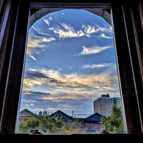Looking through an arched window twenty minutes after sunrise the blue sky is full of complex white and yellow clouds. Pointed roofs of Harlem brownstones are across the street, and a taller apartment building can be seen in the distance. The green tops of two trees are entering the frame on the bottom and right.