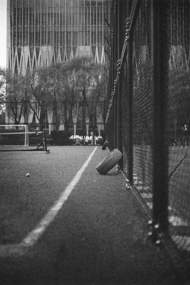 Ilford HP5 Plus 400 (FF)

English Alt Text:
A black-and-white photo of a tennis court taken from a low angle near the ground. A chain-link fence runs along the right side, with two tires leaning against it. A single tennis ball rests on the court surface. In the background, tall trees frame a striking modern building with a geometric facade made of vertical and diagonal lines. The contrast between the recreational court and the urban architecture creates a layered visual experience. The image f…