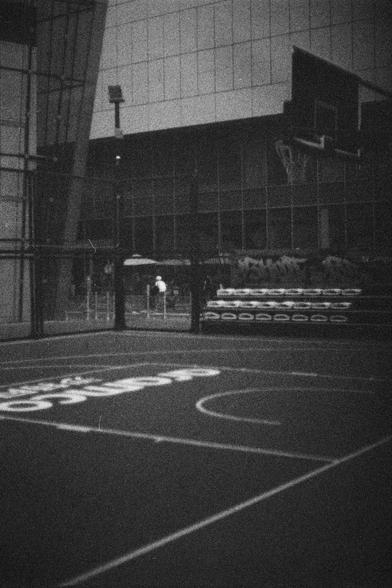 Ilford HP5 Plus 400 (FF)

English Alt Text:
A black-and-white image of an outdoor basketball court. The court is marked with clear white lines, including the three-point arc and free-throw lane. A basketball hoop with a backboard stands on the right. Behind the court, a modern building with large glass windows dominates the background. Stacked mats or barriers are visible near the fence, which encloses the court. The image has a grainy, vintage texture, evoking a nostalgic or documentary feel. …