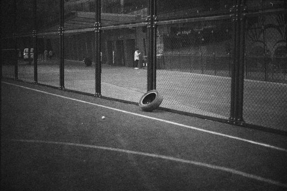 Ilford HP5 Plus 400 (FF)

English Alt Text:
A black-and-white photo of a fenced sports court, likely for tennis. A tire leans against the chain-link fence, and a ball lies nearby on the court surface. Outside the fence, a few people are walking or standing near a building. The image has a grainy texture, suggesting it was taken with a film camera or edited for a vintage look. The tire and ball inside the empty court evoke a sense of abandonment or pause, while the people outside add subtle move…