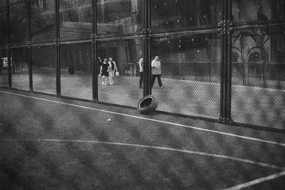 Ilford HP5 Plus 400 (FF)

English Alt Text:
A black-and-white image of a fenced sports court, possibly for tennis or basketball. Four people stand inside the court, which is enclosed by a tall chain-link fence. White boundary lines mark the ground. In the foreground, a tire leans against the fence, and a ball lies nearby. Behind the fence, a large building with architectural details looms. The monochrome tones and urban setting create a contemplative mood, blending human presence with industria…
