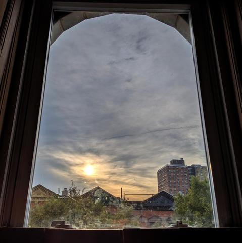 Looking through an arched window an hour after sunrise the sky is filled with dense gray-blue clouds. The sun is above the houses, slightly to the left of center, creating an orange smear above the horizon. Pointed roofs of Harlem brownstones are across the street, and a taller apartment building can be seen in the distance. The tops of two trees are on the bottom and right. The window is grimy on the bottom.