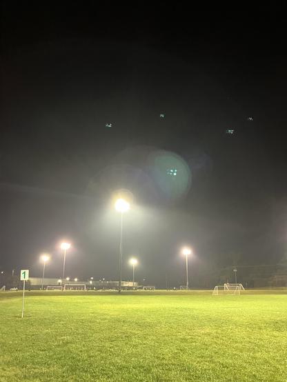 A nighttime image of a well-lit soccer field, featuring bright floodlights illuminating the grass and goalposts. A corner flag is visible in the foreground. The sky is dark, with a few faint lights visible above.