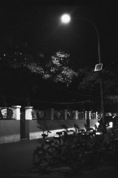 Lucky Lucky SHD 400 (FF)

English Alt Text:
A nighttime urban street scene illuminated by a bright streetlight. In the foreground, a row of identical bicycles is neatly parked, likely part of a bike-sharing system. The light casts strong shadows and highlights the metallic frames. Behind the bikes is a decorative wall with vertical bars, possibly a fence, and trees with sparse leaves visible above it. A solar panel is mounted on the streetlight pole. On the right edge, a car with headlights on …