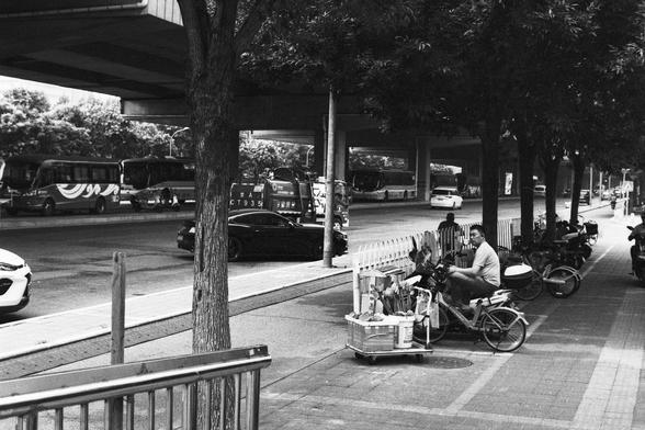 Lucky Lucky SHD 400 (FF)

English Alt Text:
A busy urban street scene under an elevated highway. On the sidewalk, a man sits on a motorcycle next to a cart filled with goods, possibly for sale. Parked motorcycles and bicycles line the pavement. Trees and a metal railing frame the foreground. In the background, multiple buses and cars, including a sleek black sports car, move along the road. The scene captures the energy of city life, blending transit, commerce, and infrastructure.

中文替代文本：
这是一个…