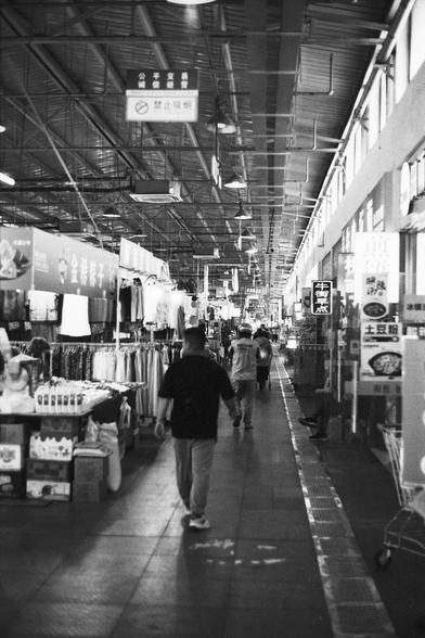 Lucky Lucky SHD 400 (FF)

English Alt Text:
A bustling indoor market corridor stretches into the distance. Shops line both sides, selling clothes, packaged goods, and miscellaneous items. Shoppers walk through, some browsing. The ceiling has exposed beams and hanging lights. Signs with Chinese characters hang above the stalls. On the right, large windows let in natural light, and posters and advertisements decorate the walls. The black-and-white palette gives the scene a documentary-style reali…