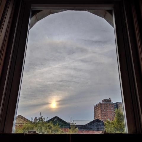 Looking through an arched window an hour after sunrise the sky is filled with dense gray-blue clouds. The yellow sun is above the houses, slightly to the left of center, obscured by the clouds. Pointed roofs of Harlem brownstones are across the street, and a taller apartment building can be seen in the distance. The tops of two trees are on the bottom and right.