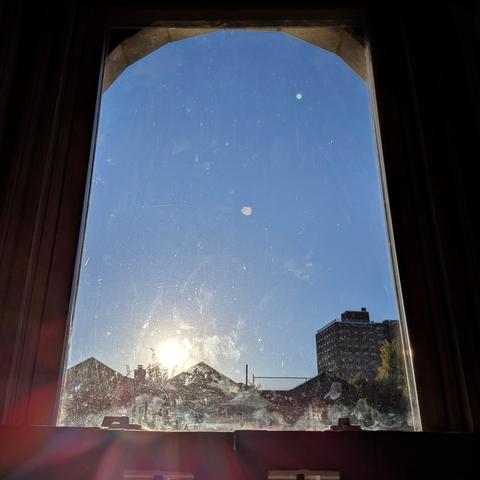 Looking through an arched window forty-five minutes after sunrise the sun is just above the buildings in the blue sky, illuminating all of the dirt and scratches on the glass, and making a lens flare with multi-colored petals on the bottom left. Pointed roofs of Harlem brownstones are silhouetted across the street, and a taller apartment building can be seen in the distance. The green tops of two trees are on the bottom and right.