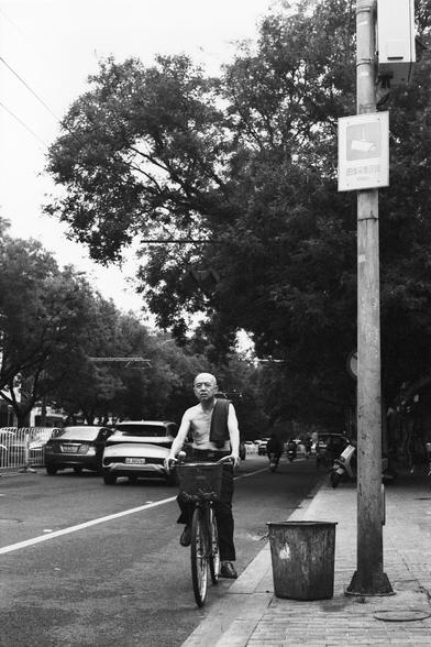 Lucky Lucky SHD 400 (FF)

English Alt Text:
A black-and-white photo shows an older shirtless man riding a bicycle on a tree-lined sidewalk. A towel hangs over one shoulder. A trash bin stands nearby. In the background, more people ride bicycles or scooters. Parked cars line the street. A utility pole displays a Chinese sign promoting public cleanliness. The scene reflects a slice of daily life in a quiet urban neighborhood.

中文替代文字：
这张黑白照片中，一位年长男子赤膊骑车在绿树成荫的人行道上，肩上搭着毛巾。旁边放着一个垃圾桶。背景中还有其他人骑着自行车或电动…