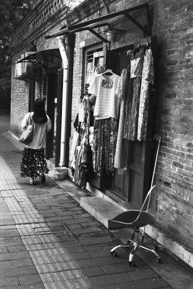 Lucky Lucky SHD 400 (FF)

English Alt Text:
A black-and-white photo captures a quiet urban street. On the right, a brick building hosts a small clothing shop with dresses and shirts hanging on the exterior wall. A red office chair with wheels sits casually on the sidewalk. A woman with long hair, wearing a patterned skirt and carrying a bag, walks away from the camera. The sidewalk is paved with bricks and includes a tactile strip for visually impaired pedestrians. The atmosphere blends traditi…