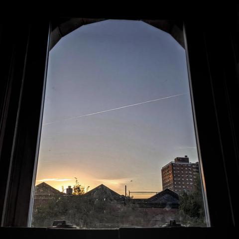 Looking through an arched window eighteen minutes after sunrise the pale blue sky is marked with bright orange clouds above the horizon, and a sharp jet trail is creating a diagonal white line across the center of the image. Pointed roofs of Harlem brownstones are silhouetted across the street, and a taller apartment building can be seen in the distance. 