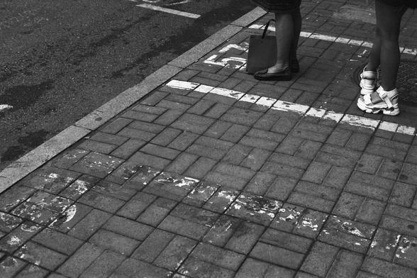 Lucky Lucky SHD 400 (FF)

English Alt Text:
A black-and-white photo of a sidewalk made of interlocking bricks. Two people stand near the edge—one wearing flat shoes and holding a shopping bag, the other in athletic shoes. White painted lines and symbols mark the ground. The scene suggests a waiting zone or designated space. The contrast between the shoes and the markings adds a sense of everyday urban life.

中文替代文字：
这是一张黑白照片，展示了由嵌砖铺成的人行道。两人站在边缘处，一人穿着平底鞋并提着购物袋，另一人穿运动鞋。地面上有白色线条和符号。画面可能是一个等候区或指定区域…