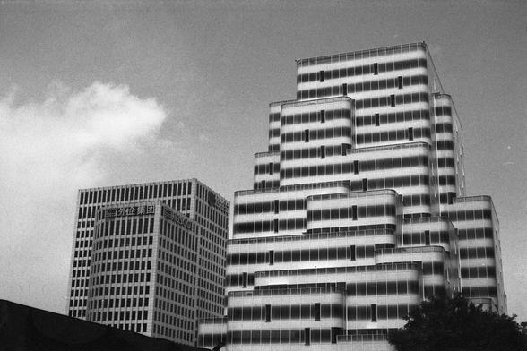 Lucky Lucky SHD 400 (FF)

English Alt Text:
A black-and-white photo of two high-rise buildings. The building on the right features a tiered design with levels that step back as they rise, creating a geometric pattern. Its facade has horizontal lines and large windows. The building on the left is more traditional, with a grid-like window layout and Chinese characters on its exterior. A tree and dark structure appear at the bottom, contrasting with the modern architecture above.

中文替代文字：
这是一张黑白照片…