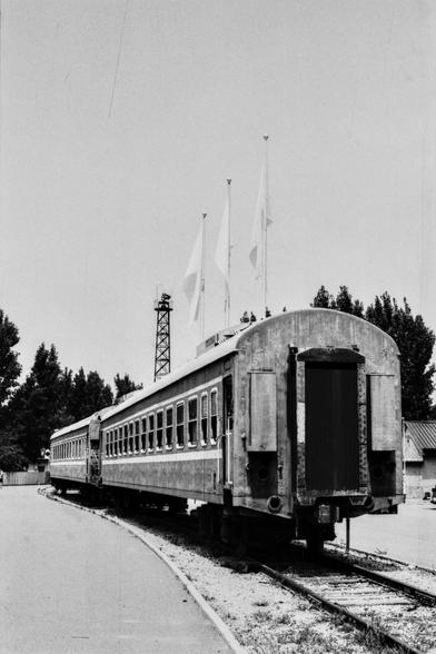 Lucky Lucky SHD 400 (FF)

English Alt Text:
A black-and-white photo of a stationary train on a curved track. The train cars are old and weathered, lined with rectangular windows. Three tall flagpoles rise above the train, each flying a flag in the breeze. Behind the train stands a metal lattice tower, possibly for communication or lighting. Trees form a natural backdrop, and a small building with a sloped roof sits nearby. The platform is quiet and empty, evoking a sense of nostalgia and stilln…