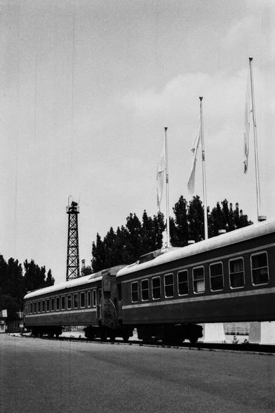 Lucky Lucky SHD 400 (FF)

English Alt Text:
A black-and-white image of a train parked at a quiet platform. The train consists of several passenger cars with visible doors and windows. Above the train, multiple tall flagpoles hold flags fluttering gently. A tall metal tower stands in the background, surrounded by trees. The platform is paved and completely empty, with no people or movement. The photo’s vintage tone suggests it was taken decades ago, evoking a sense of quiet reflection and histor…